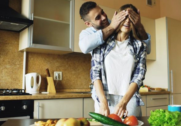 Young man covering girlfriends eyes with hands and surprising her in the kitchen at home. Loving couple cooking breakfast in the morning