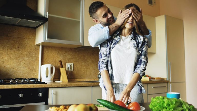 Young man covering girlfriends eyes with hands and surprising her in the kitchen at home. Loving couple cooking breakfast in the morning